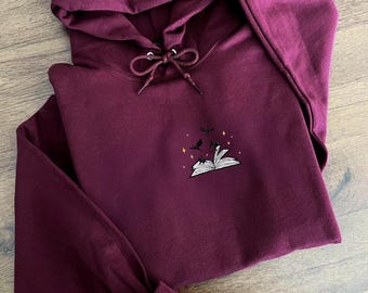 Puede incluir: Una sudadera con capucha color granate con un diseño bordado de un libro abierto con pájaros volando fuera de él. El libro está rodeado de pequeñas estrellas.