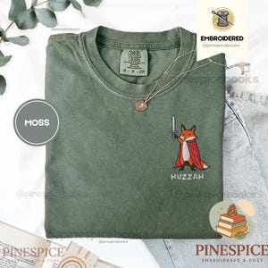Op de afbeelding: Een mosgroen t-shirt met ronde hals en een geborduurde afbeelding van een vos die een rode cape draagt en een zilveren zwaard vasthoudt. Onder de vos is het woord HUZZAH in het wit geborduurd. Het shirt heeft een Comfort Colors label.