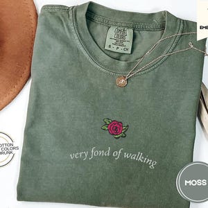 Puede incluir: Camiseta verde musgo con una rosa roja bordada y la frase "very fond of walking". La camiseta es 100% algod&oacute;n y est&aacute; preencogida. Un collar dorado con un colgante descansa sobre la camiseta.
