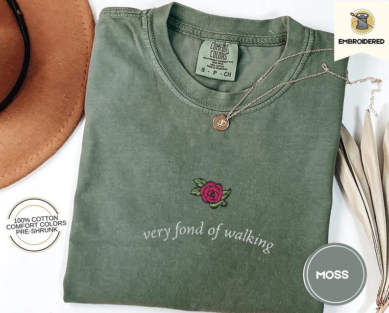 Puede incluir: Camiseta verde musgo con una rosa roja bordada y la frase "very fond of walking". La camiseta es 100% algod&oacute;n y est&aacute; preencogida. Un collar dorado con un colgante descansa sobre la camiseta.