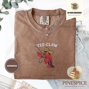 May include: Espresso-colored t-shirt with the embroidered phrase "Yee-Claw" and a cartoon lobster wearing a cowboy hat. The shirt has a crew neck and short sleeves. A silver necklace with a pendant is draped on the shirt.