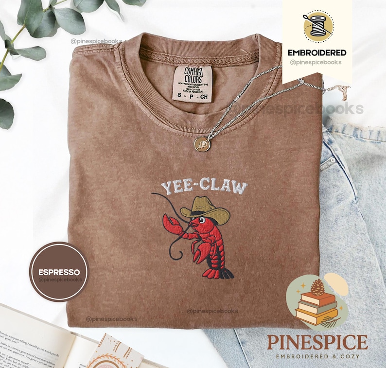 May include: Espresso-colored t-shirt with the embroidered phrase "Yee-Claw" and a cartoon lobster wearing a cowboy hat. The shirt has a crew neck and short sleeves. A silver necklace with a pendant is draped on the shirt.
