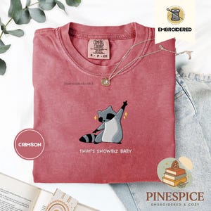 Könnte beinhalten: Karmesinrotes T-Shirt mit einem gestickten Waschbären-Design. Der Waschbär ist in Grau und Weiß dargestellt, mit einem schwarz gestreiften Schwanz, der posiert. Der Text "THAT'S SHOWBIZ BABY" steht darunter. Das Shirt hat einen Rundhalsausschnitt und kurze Ärmel.