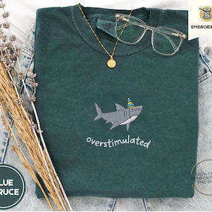 May include: A dark teal, folded t-shirt with an embroidered shark wearing a party hat. The text "overstimulated" is below the shark. A gold necklace, clear glasses, and dried plants are on the shirt. The shirt is labeled "Blue Spruce".