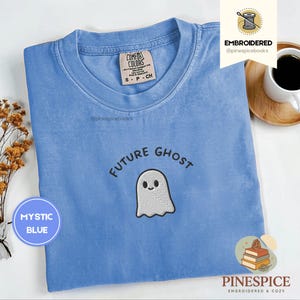 May include: Mystic blue t-shirt with the embroidered text "FUTURE GHOST" and a smiling ghost graphic. The shirt has a crew neck and a Comfort Colors tag. The brand name "PINESPICE" is at the bottom.