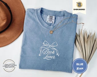 Peut inclure: Un t-shirt bleu jean avec une broderie blanche d'un nœud et les mots "Book Lover".