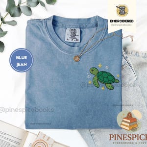 May include: A blue jean colored t-shirt featuring an embroidered green sea turtle on the chest. The shirt has a crew neckline and a Comfort Colors label inside the collar. Small yellow star icons and white bubbles surround the turtle design.