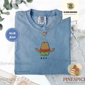 Puede incluir: Una camiseta color azul jean con un bordado de una criatura verde que lleva un sombrero de vaquero marrón con una banda roja. Debajo de la criatura, el texto yee está bordado en negro. La camiseta tiene una etiqueta de la marca Comfort Colors.
