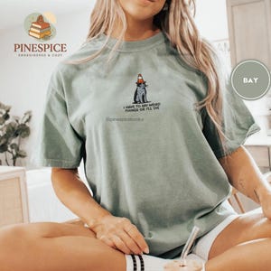 May include: Sage green t-shirt with a graphic of a cat wearing a traffic cone hat. The text "I HAVE TO SAY WEIRD THINGS OR I'LL DIE" is below the cat. The shirt also has the text "PINESPICE EMBROIDERED & COZY" and "@pinespicebooks".