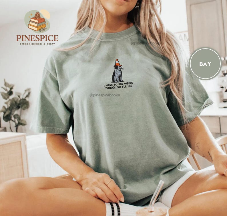 May include: Sage green t-shirt with a graphic of a cat wearing a traffic cone hat. The text "I HAVE TO SAY WEIRD THINGS OR I'LL DIE" is below the cat. The shirt also has the text "PINESPICE EMBROIDERED & COZY" and "@pinespicebooks".