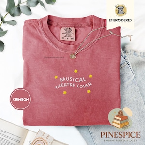 Könnte beinhalten: Ein karmesinrotes Kurzarm-T-Shirt mit Rundhalsausschnitt. Das Shirt zeigt den weiß gestickten Schriftzug "MUSICAL THEATRE LOVER", umgeben von fünf kleinen gelben gestickten Sternen. Ein Etikett gibt die Größe S an.