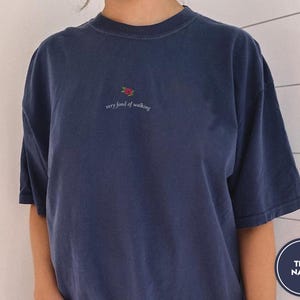 Puede incluir: Camiseta azul marino con una peque&ntilde;a rosa bordada y la frase "very fond of walking". La camiseta tiene mangas cortas y un corte relajado. El color se describe como "True Navy".