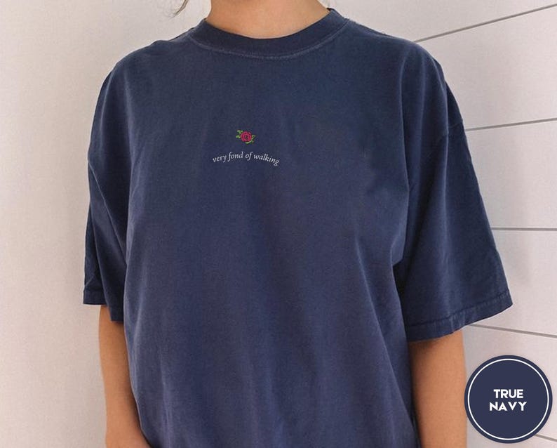 Puede incluir: Camiseta azul marino con una peque&ntilde;a rosa bordada y la frase "very fond of walking". La camiseta tiene mangas cortas y un corte relajado. El color se describe como "True Navy".