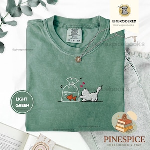 Puede incluir: Una camiseta verde claro con un diseño bordado de un gato blanco que intenta alcanzar a un pez dorado en una bolsa de plástico. El gato está situado a la derecha de la bolsa, con dos pequeños corazones rojos flotando sobre su espalda.