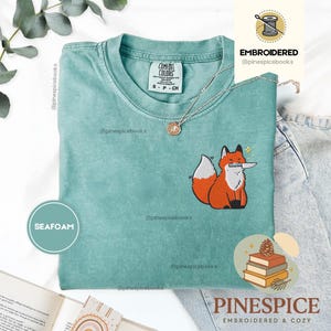 Può includere: Una t-shirt a maniche corte color verde schiuma di mare con un ricamo raffigurante una volpe arancione che tiene un coltello argentato in bocca. La maglia presenta un girocollo e un'etichetta Comfort Colors all'interno. Una collana argentata poggia sul tessuto.