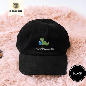 May include: Black corduroy baseball cap with an embroidered green bookworm design, wearing glasses and holding a blue book. The word "bookworm" is embroidered below the worm. The cap is on a pink, fluffy surface.