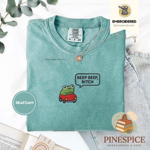 Könnte beinhalten: Türkisgrünes T-Shirt mit einer gestickten Froschfigur, die ein rotes Auto fährt, und dem Text "BEEP BEEP, BITCH" in einer Sprechblase. Das Shirt hat einen Rundhalsausschnitt und kurze Ärmel. Eine silberne Halskette mit Anhänger liegt auf dem Shirt.