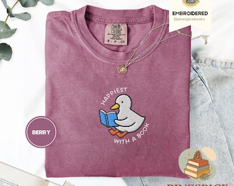 Peut inclure: T-shirt Comfort Colors de couleur baie avec une broderie d'un canard lisant un livre. Le texte "Happiest with a Book" est brodé autour du canard. Un collier doré et le texte "EMBROIDERED @pinespicebooks" sont également visibles.