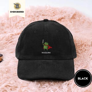 Könnte beinhalten: Schwarze Cord-Baseballkappe mit einem gestickten Design einer grünen Froschfigur mit rotem Umhang und Schwert. Das Wort "Huzzah" ist darunter gestickt. Die Kappe hat einen gebogenen Schirm und einen verstellbaren Riemen.
