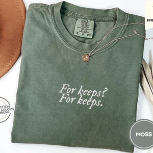 Puede incluir: Una camiseta verde musgo con el texto bordado "For keeps? For keeps." Un collar dorado con un colgante descansa sobre la camisa. La camiseta tiene cuello redondo y mangas cortas. La camiseta es 100% algodón.