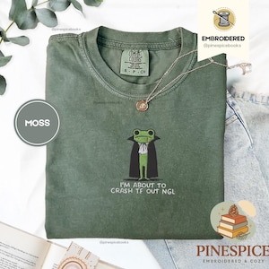 Puede incluir: Camiseta verde musgo con un bordado de una rana vestida de vampiro, con el texto "I'M ABOUT TO CRASH TF OUT NGL". La camiseta tiene cuello redondo y mangas cortas. Un collar dorado está drapeado sobre la camiseta.