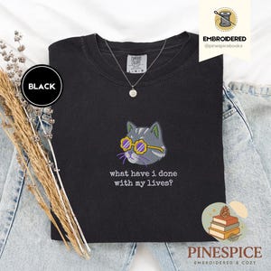 May include: Black Comfort Colors sweatshirt with an embroidered cat wearing yellow sunglasses. The cat is gray with green accents. The text "what have I done with my lives?" is below the cat.