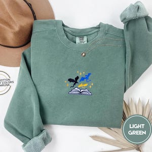 May include: A light green sweatshirt with an embroidered design of three dragons flying around a book. The text "100% COTTON COMFORT COLORS PRE-SHRUNK" is visible on the left side of the image. The text "LIGHT GREEN" is visible on the right side of the image.