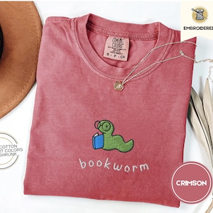 May include: A crimson red t-shirt with a green worm holding a book and the text "bookworm" embroidered on the front. The shirt is made of 100% cotton and is pre-shrunk.