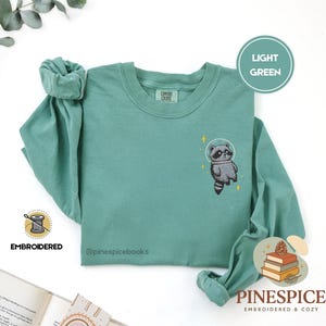 May include: Light green long-sleeved shirt featuring an embroidered raccoon in a space helmet. The shirt has the text "Comfort Colors" on the collar and "EMBROIDERED" near the sleeve. The brand name "PINESPICE" is also visible.
