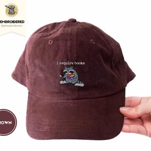 May include: Brown corduroy baseball cap with an embroidered raccoon holding books. The text "i require books" is above the raccoon. The cap has a curved brim and a button on top.