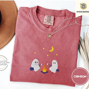 May include: A crimson red t-shirt with an embroidered design of two ghosts roasting marshmallows over a campfire. The text "COMFORT COLORS" and "S-P-CH" is visible on the shirt. The shirt is 100% cotton, comfort colors, and pre-shrunk.