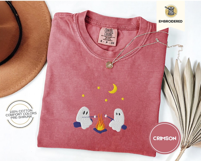 May include: A crimson red t-shirt with an embroidered design of two ghosts roasting marshmallows over a campfire. The text "COMFORT COLORS" and "S-P-CH" is visible on the shirt. The shirt is 100% cotton, comfort colors, and pre-shrunk.
