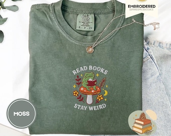 Könnte beinhalten: Moosgrünes T-Shirt mit gesticktem Design. Das Design zeigt einen Frosch, der ein Buch auf einem Pilz liest, mit dem Text "READ BOOKS STAY WEIRD". Eine silberne Halskette mit Anhänger befindet sich auf dem Shirt. Das Shirt ist gefaltet.