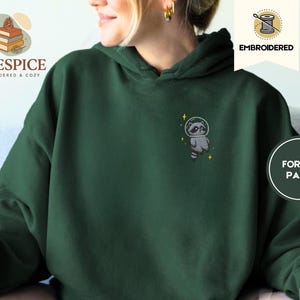 K&ouml;nnte beinhalten: Dunkelgr&uuml;ner Kapuzenpullover mit gesticktem Waschb&auml;ren im Weltraumanzug. Der Waschb&auml;r ist grau und wei&szlig; mit einem schwarzen Helm und gelben Sternen. Der Hoodie hat eine lockere Passform.