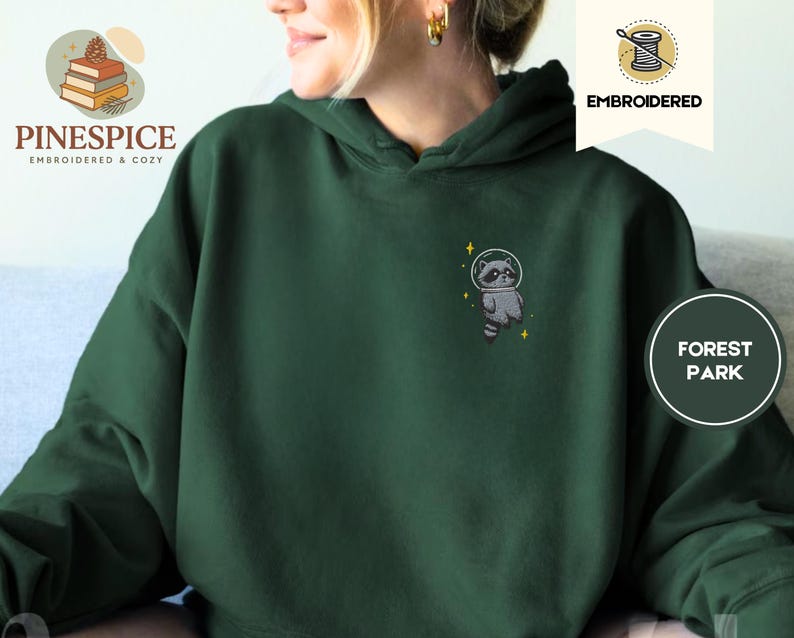 K&ouml;nnte beinhalten: Dunkelgr&uuml;ner Kapuzenpullover mit gesticktem Waschb&auml;ren im Weltraumanzug. Der Waschb&auml;r ist grau und wei&szlig; mit einem schwarzen Helm und gelben Sternen. Der Hoodie hat eine lockere Passform.