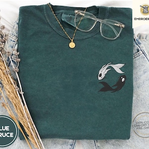 May include: A dark green t-shirt with an embroidered design of two fish, one white and one black, in a yin-yang style. The shirt is made of 100% cotton and is pre-shrunk. The text "EMBROIDERED" is visible in the top right corner of the image.