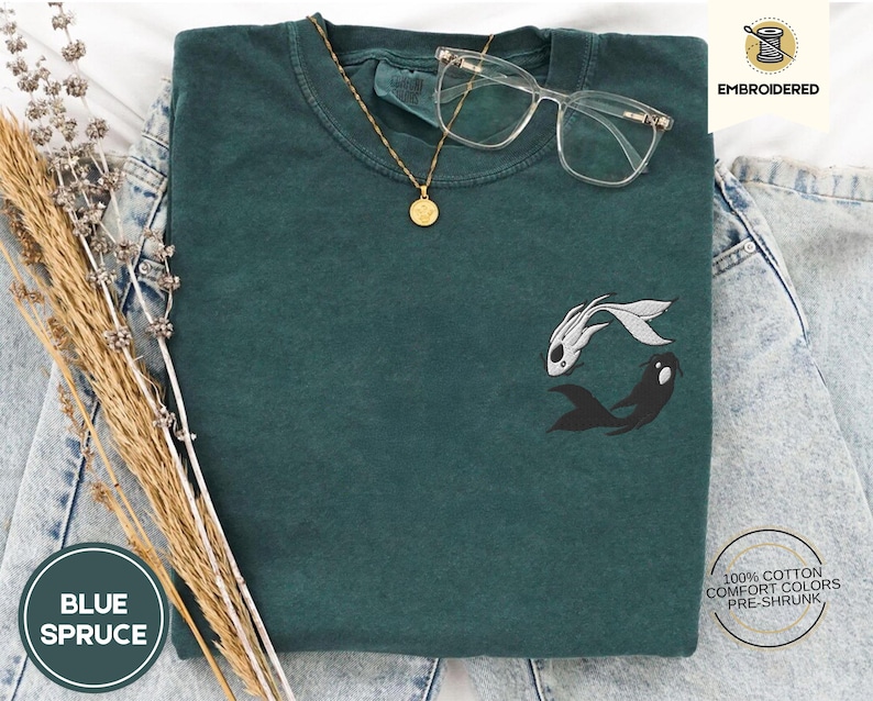 May include: A dark green t-shirt with an embroidered design of two fish, one white and one black, in a yin-yang style. The shirt is made of 100% cotton and is pre-shrunk. The text "EMBROIDERED" is visible in the top right corner of the image.