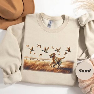 May include: Sand-colored sweatshirt featuring a Labrador Retriever running through a field with flying ducks. The design includes a brown and tan color palette, with a pumpkin and hat in the background. The word "Sand" is printed on a circular label.