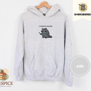 May include: Ash-colored hoodie with a kangaroo pocket and drawstring hood. The embroidered design features a raccoon holding a knife, with the text "I require snacks" above it. The hoodie is displayed on a wooden hanger.