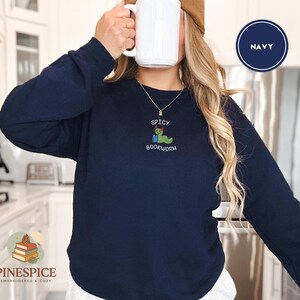 Puede incluir: Sudadera azul marino con dise&ntilde;o bordado "Spicy Bookworm". El dise&ntilde;o presenta un gusano verde con un chile rojo. La sudadera es de manga larga y cuello redondo.