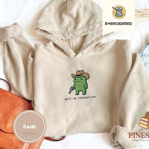 Può includere: Una felpa con cappuccio color sabbia con un ricamo raffigurante una rana verde che indossa un cappello da cowboy marrone e tiene in mano un piccolo revolver argentato. Sotto la rana, il testo WOT IN TERNATION è ricamato in nero.
