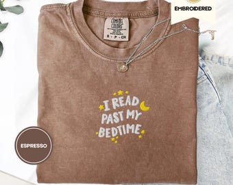 Peut inclure: T-shirt couleur expresso avec la phrase brodée "I READ PAST MY BEDTIME" en jaune et blanc. Le t-shirt a un col rond et des manches courtes. Un collier argenté avec un pendentif est drapé sur le t-shirt.