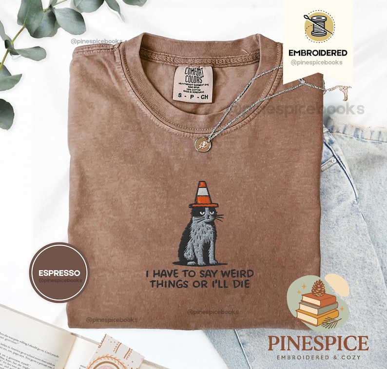 May include: Espresso-colored t-shirt with a graphic of a cat wearing a traffic cone hat. The text "I HAVE TO SAY WEIRD THINGS OR I'LL DIE" is below the cat. The shirt has a crew neck and short sleeves. A silver necklace with a pendant is on the shirt.
