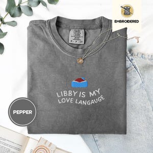 以下が含まれることがあります： 「Libby is my love language」の刺繍と小さな家のデザインが施されたグレーのComfort Colors Tシャツ。 シャツのサイズはSで、丸いネックラインです。 ペンダント付きのシルバーネックレスも表示されています。