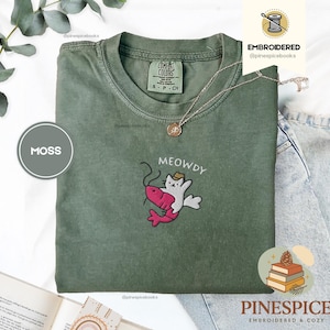 Può includere: T-shirt verde muschio con un disegno ricamato di un gatto con un cappello che cavalca un gambero rosa, con la scritta "MEOWDY" sopra. La maglietta ha un'etichetta Comfort Colors e una collana d'oro. Taglia S.