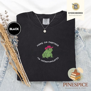 Puede incluir: Camiseta negra Comfort Colors con un diseño bordado que presenta una rana verde con un sombrero de vaquero rosa. El texto dice "Hang on Partner, I'm Overstimulated". Se ve un collar plateado.