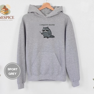 May include: Sport grey hoodie with a kangaroo pocket and drawstring hood. The front features an embroidered raccoon holding a knife, with the text "I require snacks" above it.
