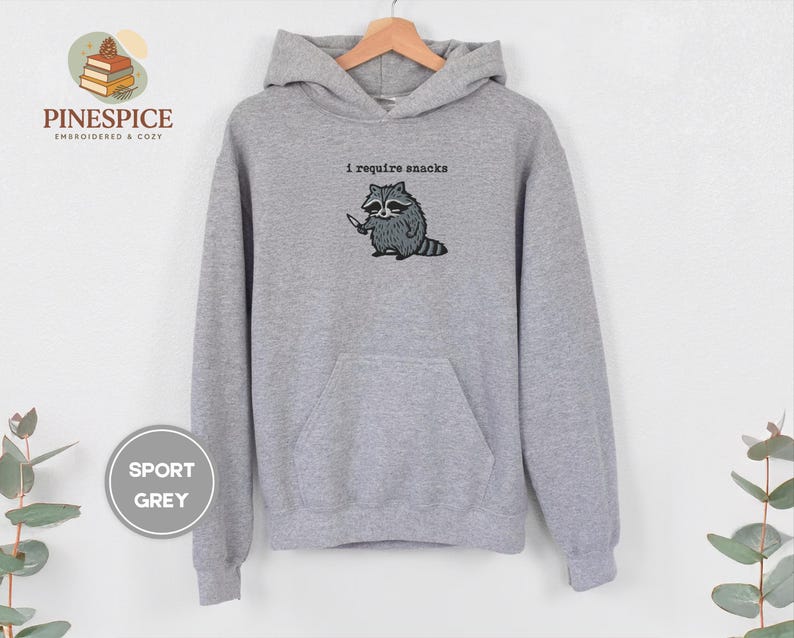 May include: Sport grey hoodie with a kangaroo pocket and drawstring hood. The front features an embroidered raccoon holding a knife, with the text "I require snacks" above it.
