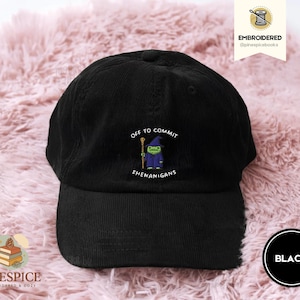 May include: Black corduroy baseball cap with the embroidered phrase "Off to Commit Shenanigans" and a cartoon wizard. The cap has a curved brim and a black adjustable strap. The cap is on a pink, fluffy surface.