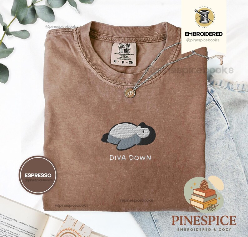 May include: Espresso-coloured t-shirt with a crew neck and short sleeves. The shirt features an embroidered design of a sleeping creature with the text "DIVA DOWN". A silver necklace with a pendant is visible. The shirt is from the brand Comfort Colors.